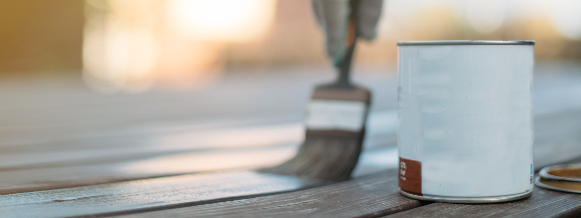 A hand paints a wooden floor and a paint can lies to its left. 