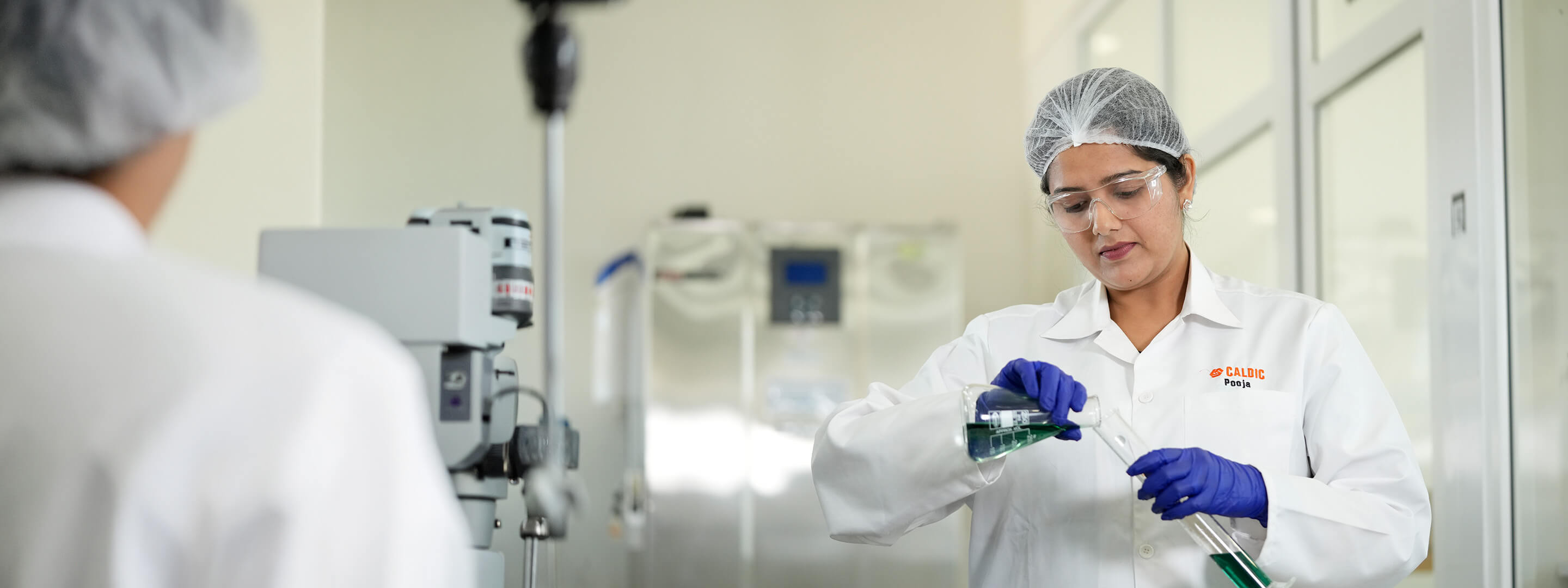 Laboratory professional pouring green liquid between glass flasks during ingredient formulation process